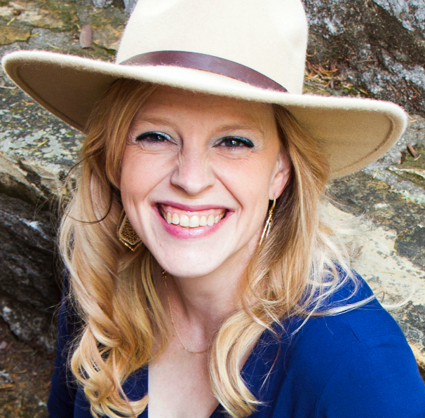 Lainey La Shay Headshot Lainey La Shay smiling brightly in an outdoor setting, wearing a wide-brimmed beige hat with a brown band and a deep blue top.